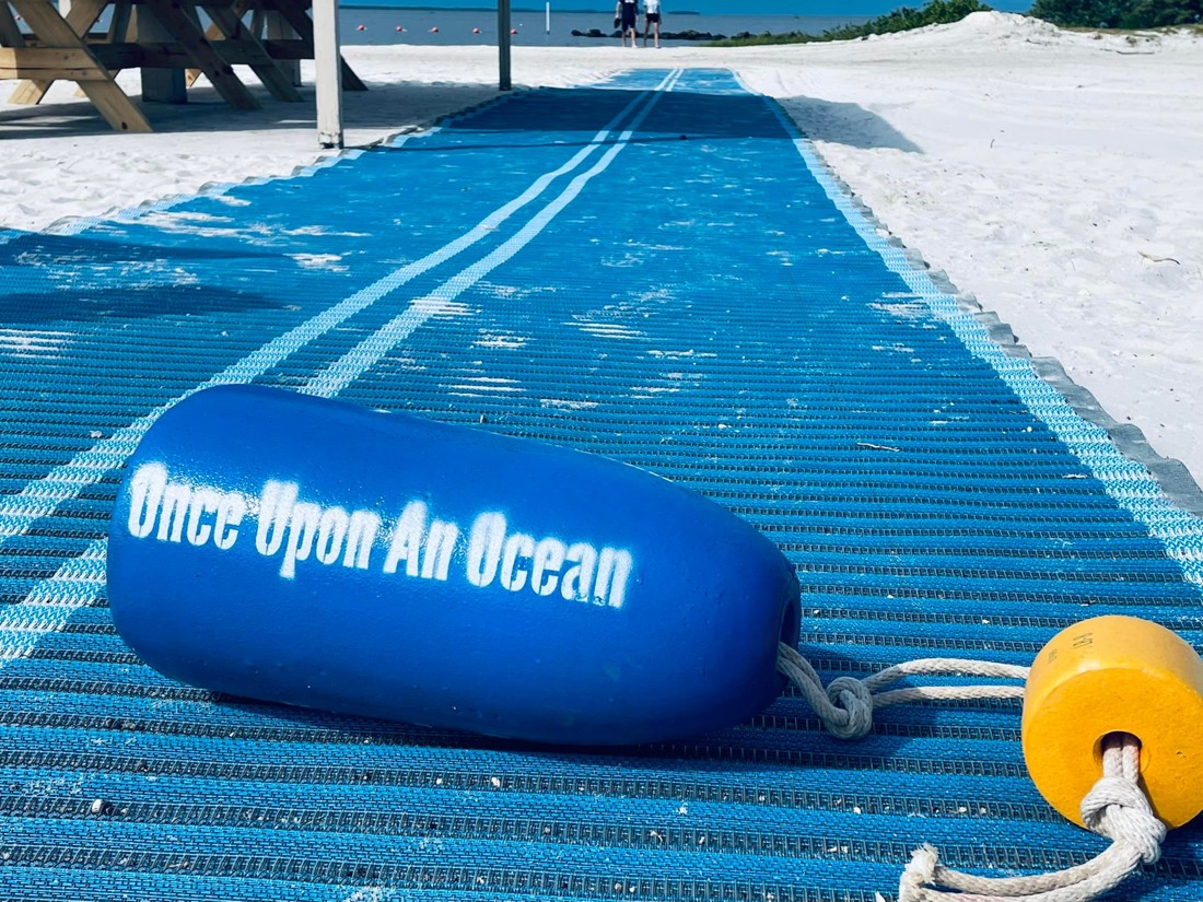A blue Once Upon an Ocean float beside a beach access mat on the sand.